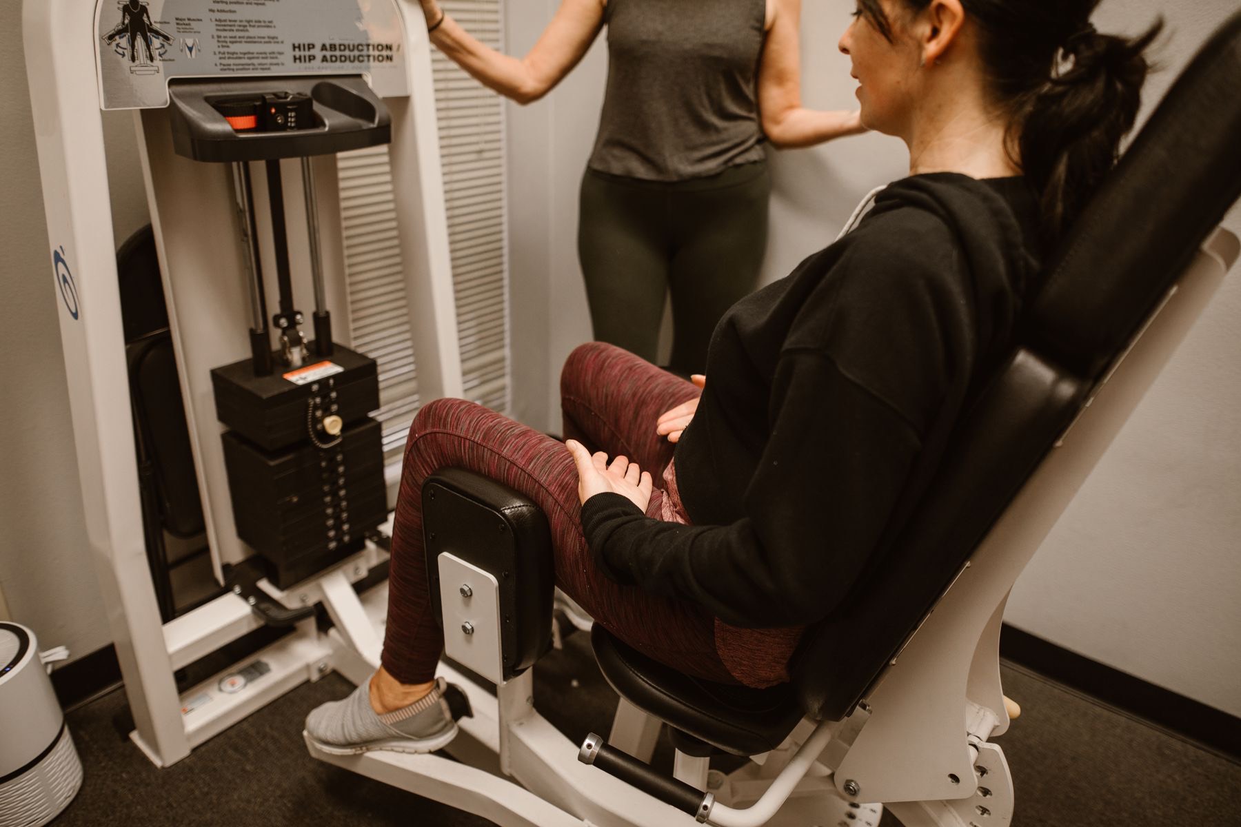 A client seated on a hip abduction and adduction machine with a trainer nearby